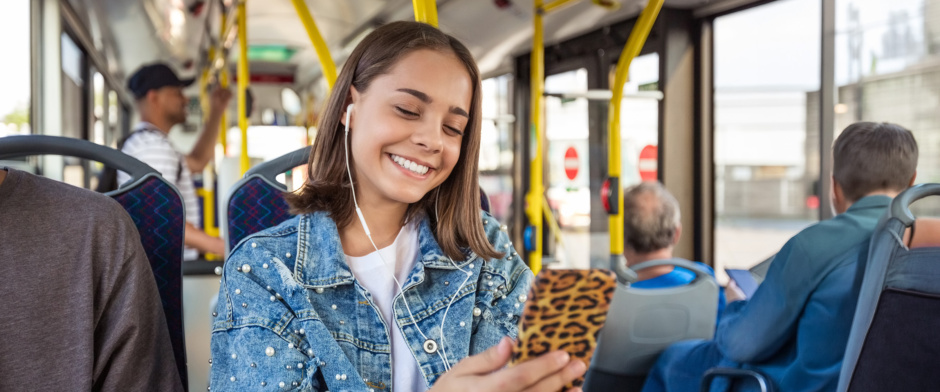 Eine Jugendliche fährt in einem Bus des öffentlichen Nahverkehrs. Sie schaut auf ihr Smartphone und lächelt.