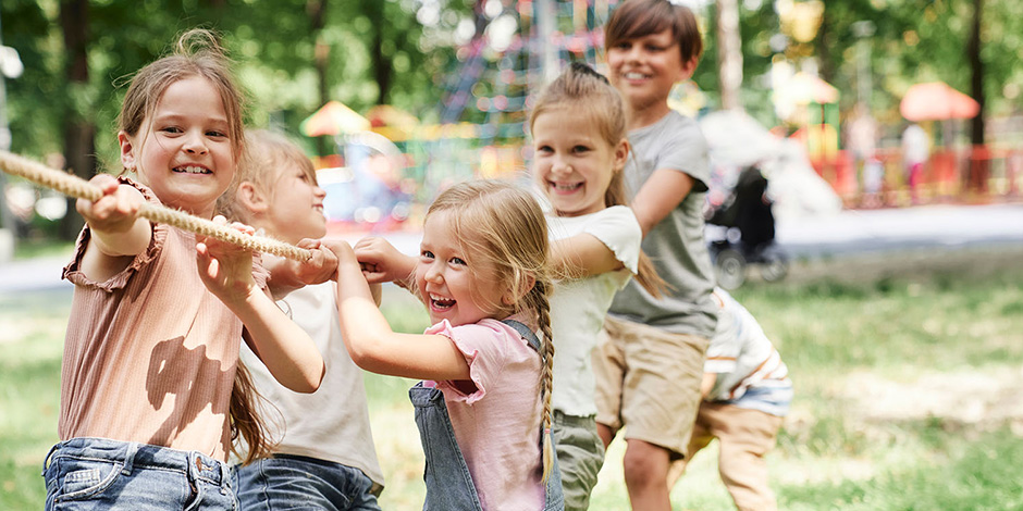 Kinder spielen in einem Park Tauziehen. Im Hintergrund ist ein Spielplatz zu sehen.